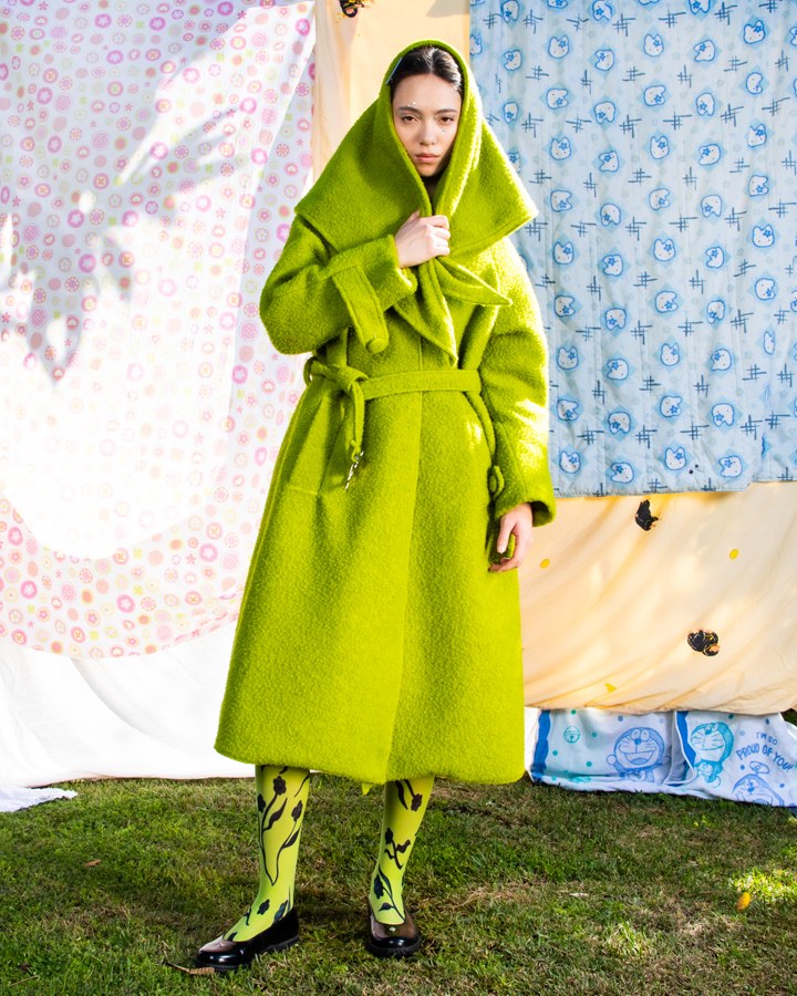 Full body image of a female model in a green matching coat and scarf set against a backdrop of patterned quilts.